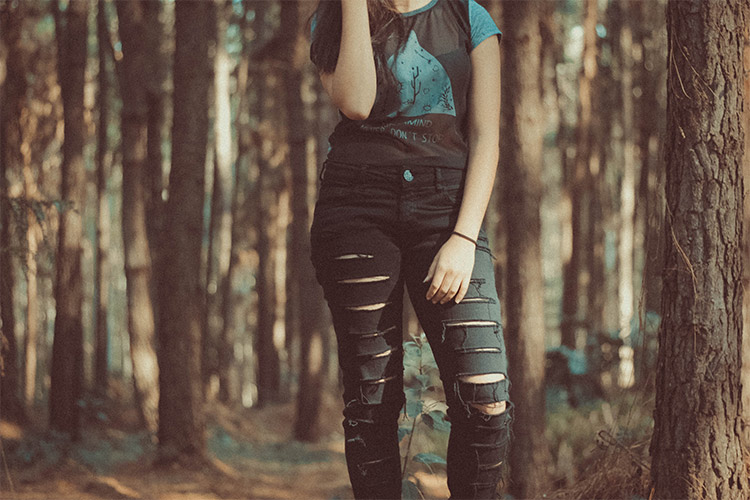 Una chica vistiendo camiseta negra, manga corta y azul, estampado azul. Además un pantalón negro modelo desgastado. Ella posa para la fotografía y se encuentra en un monte. 