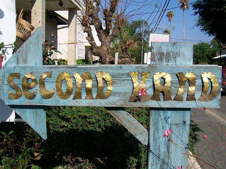 Un cartel artesano de madera azul con forma de flecha y que pone “second hand”, indica que a la izquierda habrá ropa de segunda mano. 