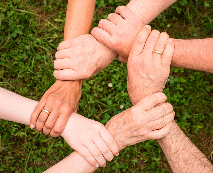 Seis manos de personas de diferentes edades, razas y sexo, agarrándose en cadena, representando apoyo humanitario.