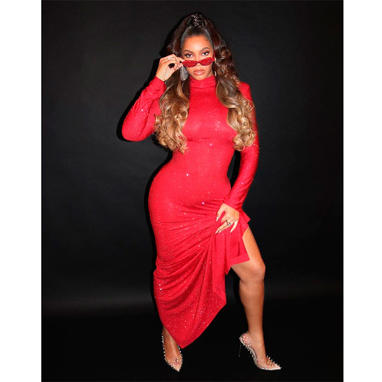 La famosa cantante Beyoncé posa con con un vestido largo rojo con brillantes y la pierna al descubierto. Lo combina con unos zapatos de tacón transparentes con tachas, mostrando lo que podría ser su look outfit Navidad