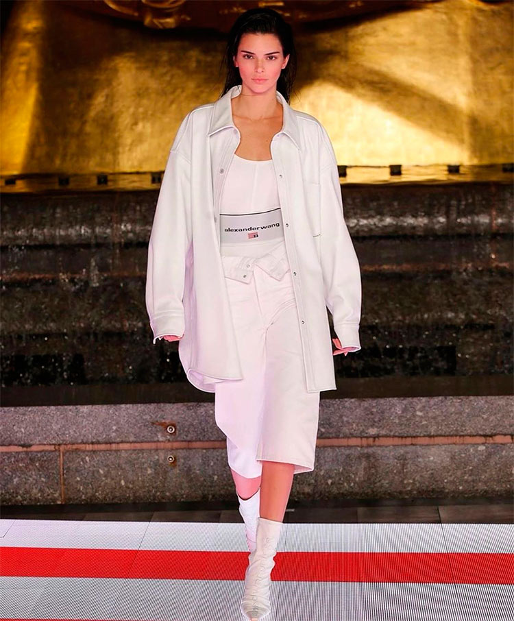Kendall Jenner desfila con un look camisa blanca oversize abierta con un top blanco debajo, unos pantalones a la altura de la rodilla y unos botines, un total white look.