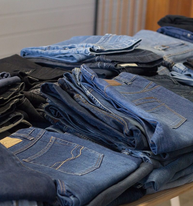 Pantalones vaqueros de colores azules, grises y negros, prenda que causa gran contaminación medioambiente, colocados sobre una balda de madera en una tienda de ropa.