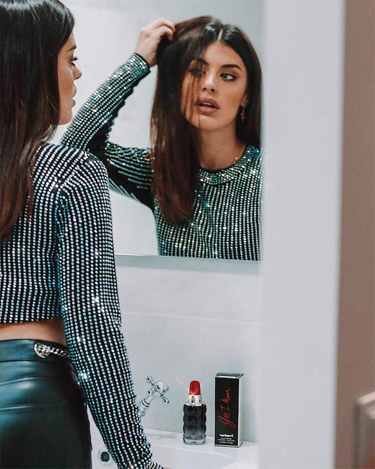 Aparece la top 1 influencer España, Dulceida, posando en delante de un espejo con un outfit de fiesta negro y plateado, en el lavabo está posada una colonia que ella publicita.