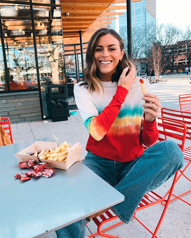 La influencer Claire Guentz posa comiendo patatas alegre, con un pantalón vaquero y una sudadera tie-dye de varios colores, completando un outfit tendencia temporada 2020.