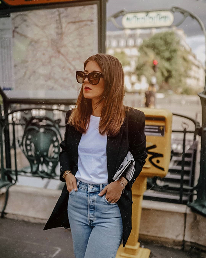 Elodie in Paris posando en la calle vestida con camiseta blanca, vaqueros azules, chaqueta negra y gafas de sol, usando truco moda.
