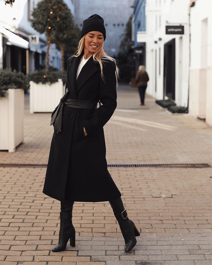 Marta Carriedo vestida entera de negro, con un abrigo largo, botas con tacón, cinturón y gorro, posando en la calle, usando un moda truco.