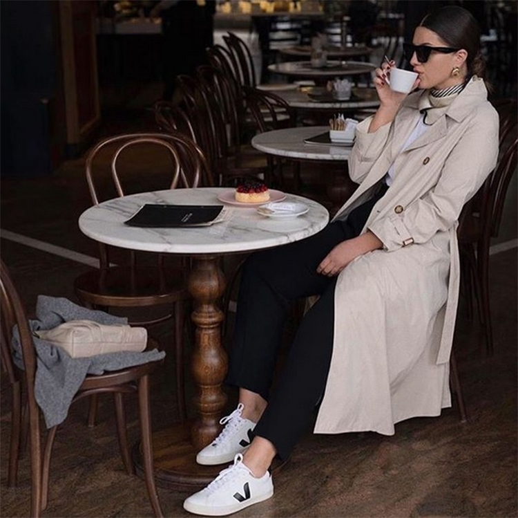 Mujer vestida con un abrigo beige, pantalones negros y unas zapatillas veganas baratas blancas, tomando café en una cafetería.