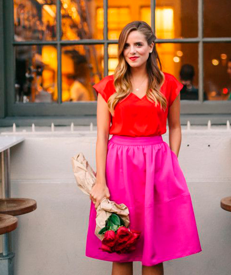 Una mujer posa con outfit San Valentín 2020 rosa y rojo con una falda midi y una camiseta, es un look mujer San Valentín ideal. Posa junto a un ramo de rosas.