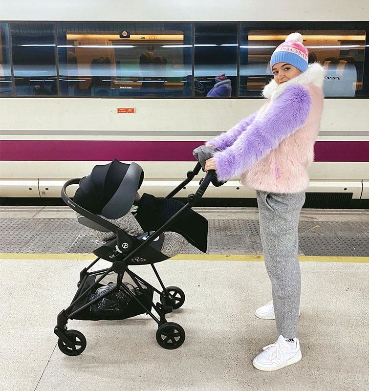 La influencer Laura Escanes posa en un andén junto al carrito de su bebé con unp de los looks invierno mujer, informal pero cool, con chándal, sneakers blancas y una chaqueta de pelo tendencia tricolor.