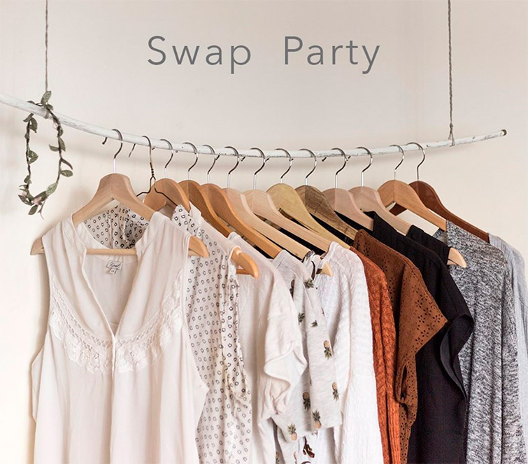 Camisetas, blusas y jerséis colgados en perchas y en una barra debajo de un cartel en la pared blanca que indica swap party