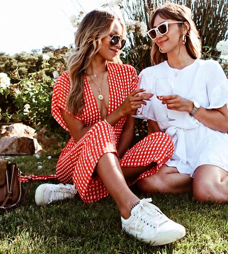 La influencer Julie Sariñana @sincerelyjules sentada en la hierba junto a una amiga, luce un outfit compuesto por vestido de cuadros vichyy zapatillas mujer blancas
