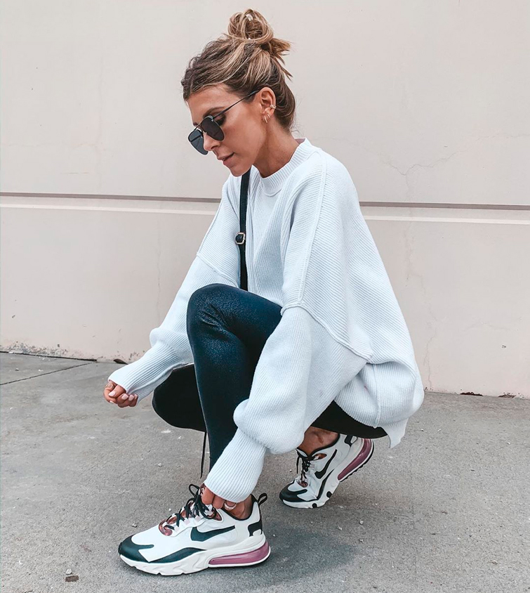 La influencer @cellajaneblog de cuclillas atándose unas zapatillas blancas mujer Nike, que combina con unos pantalones negros y un jersey blanco
