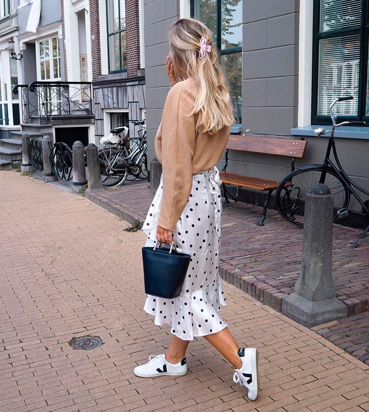 la micro influencer @julesdevrieze camina por la calle luciendo un look de falda midi con estamapdo de topos, jersey color camel y zapatillas blancas mujer Veja
