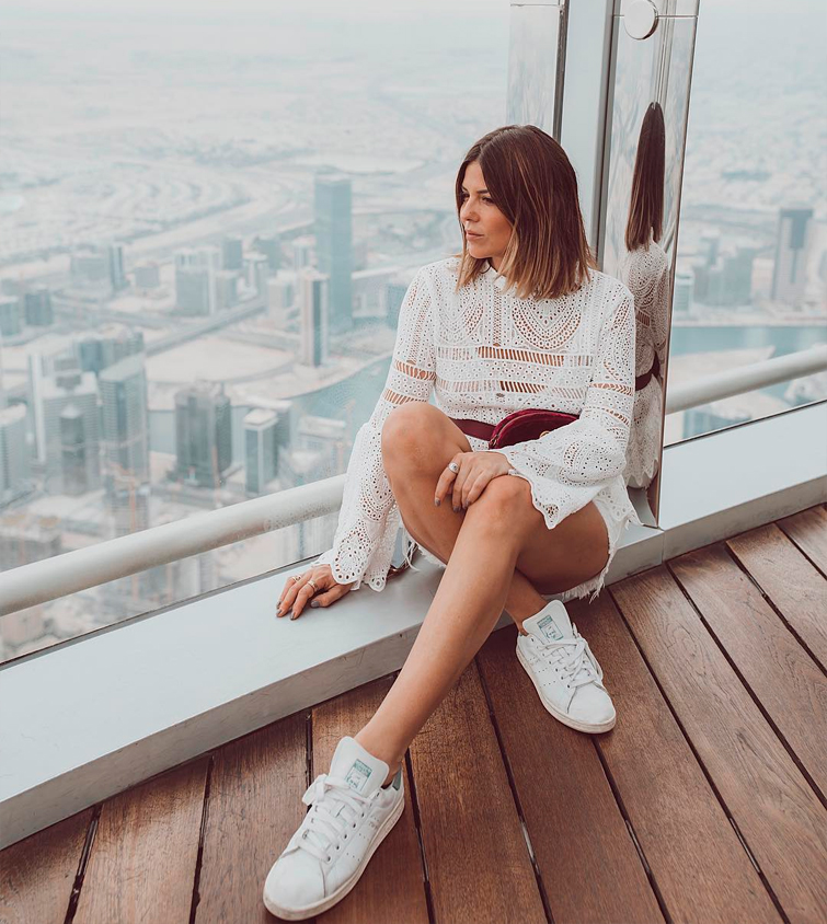 La influencer Natalia Cabezas @trendy_taste sentada en una terraza vestida con un vestido mini blanzo ibizenco y unas deportivas blancas Adidas
