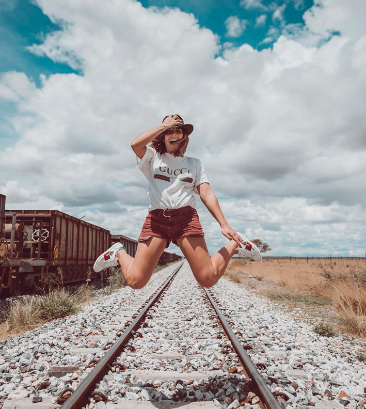 Natalia Cabezas, autora del blog de moda Trendy Taste, saltando en unas vías de tren en las que se ve que luce una camiseta blanca de Gucci, bermudas marrones y zapatillas mujer blancas
