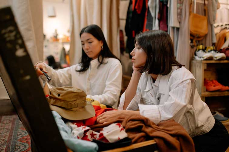 Dos mujeres jóvenes rebuscan en un baúl lleno de prendas, están en una de las tiendas que compren ropa de segunda mano en Zaragoza