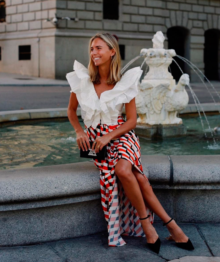 Lucía Bárcena, una de las celebridades de moda, posa sentada junto a una fuente con un conjunto de blusa blanca y falda estampada. Una de las ideas look graduación más distintas al resto
